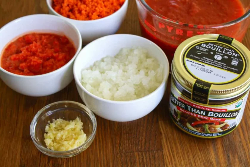 Ingredients to make fake Bolognese sauce, included mince garlic, ground onion, ground carrot, ground red bell pepper, tomatoes, all in glass and ceramic bowls, plus a jar of Better Than Bouillon, on a wooden surface.