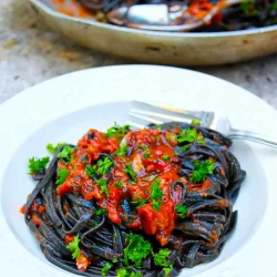 Un plato de linguini negros alla puttanesca con un poco de perejil. Al fondo se ve un sartén con más pasta.