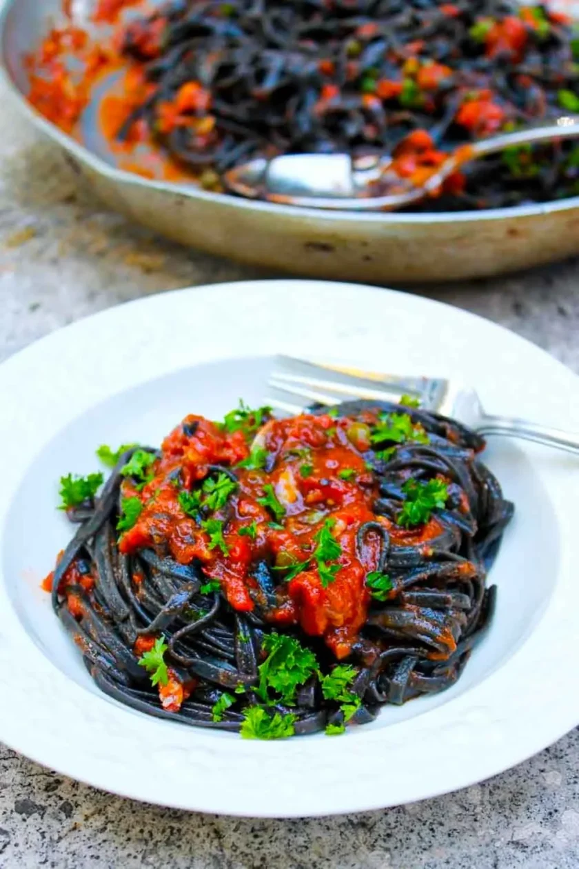 Pasta alla puttanesca - SAVOIR FAIRE by enrilemoine Un plato de linguini negros alla puttanesca con un poco de perejil. Al fondo se ve un sartén con más pasta.