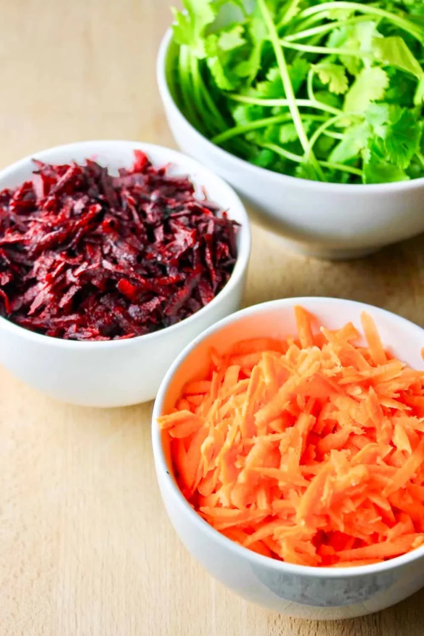  three white bowls with cilantro, grated beet and grated carrot, on a wooden surface.