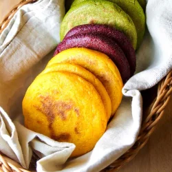 a basket with 8 veggie-colored arepas made with carrot, beet and cilantro, on a beige linen towel.