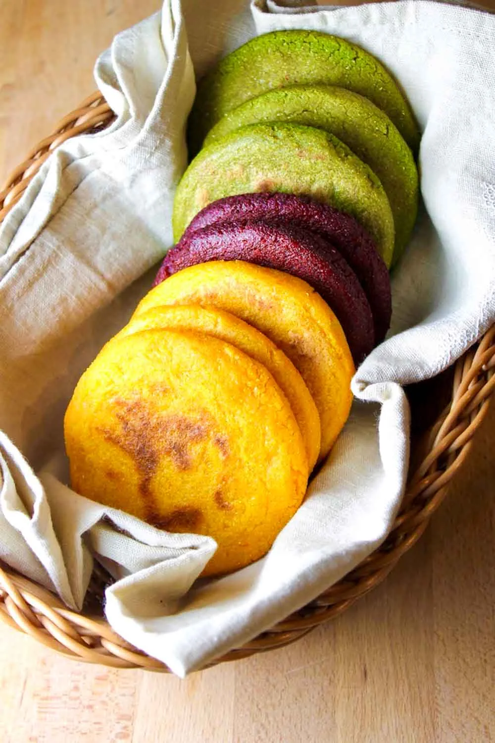 una cesta con 8 arepas multicolores de zanahoria, remolacha y cilantro, sobre una toalla de lino beige.