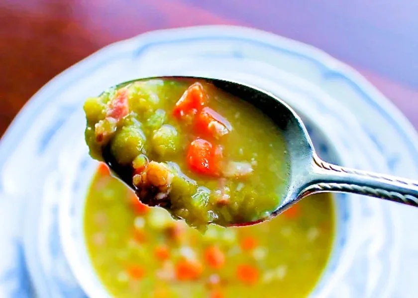 una cuchara de sopa de arvejas con jam´øn y al fon se ve el plato de sopa borroso.