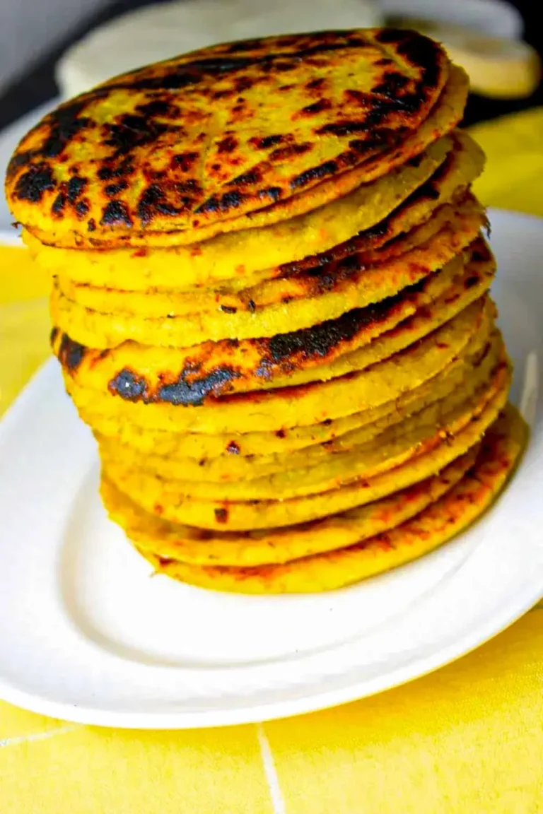 Una alta pila de tortas de maíz redondas y asadas a la parrilla descansa sobre un plato blanco, sobre un mantel amarillo.