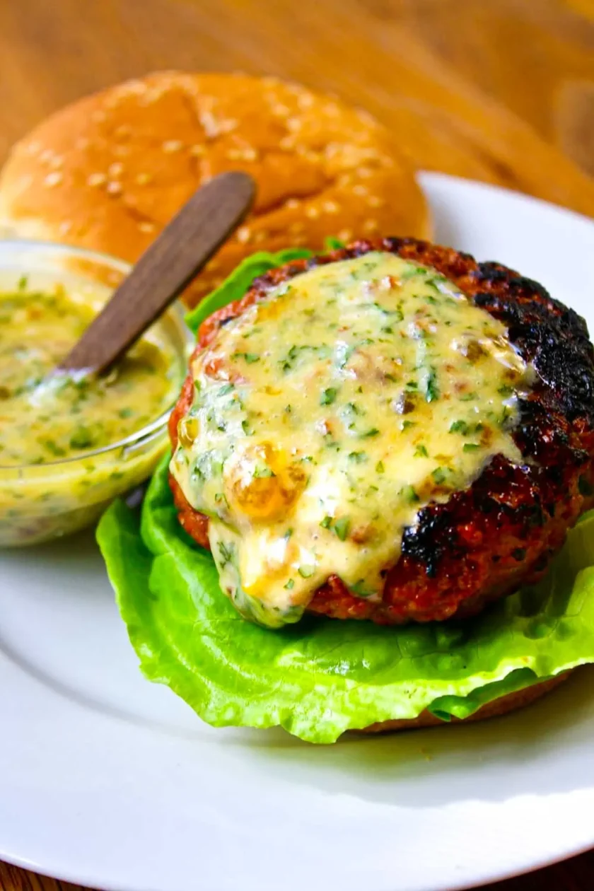 Una hamburguesa a la parrilla con mantequilla de hierbas derretida sobre lechuga, servida con un panecillo de sésamo y un pequeño cuenco de salsa de hierbas en un plato blanco.