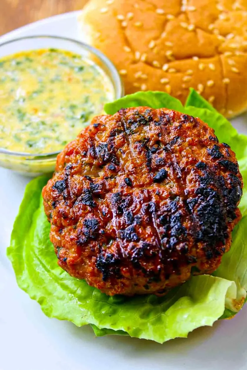 Una hamburguesa a la parrilla sobre una hoja de lechuga, con un pan con semillas de sésamo y un pequeño cuenco de salsa al fondo.
