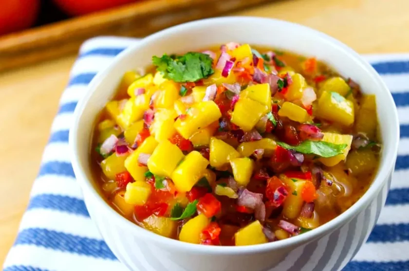 A bowl of mango ceviche with diced mango, red onion, red bell pepper, cilantro, and tomatoes resting on a blue and white striped tablecloth.