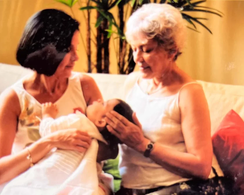 Dos mujeres sentadas en un sofá, una con un bebé en brazos mientras la otra le toca suavemente la cabeza. En el fondo hay plantas verdes.
