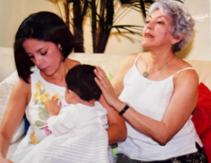 Una mujer sostiene a un bebé mientras otra mujer mayor se sienta a su lado, apoyando la mano en el hombro de la primera. Están sentadas en el interior, cerca de una planta.
