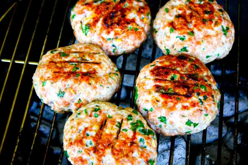 Cinco hamburguesas sazonadas con hierbas visibles se asan en una barbacoa, mostrando marcas de la parrilla y una superficie ligeramente dorada.