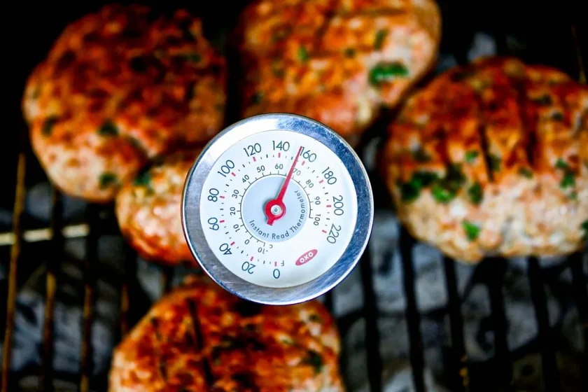 Se sostiene un termómetro para carne que marca unos 140°F sobre varias hamburguesas a la parrilla con hierbas visibles, cocinándose en una parrilla.