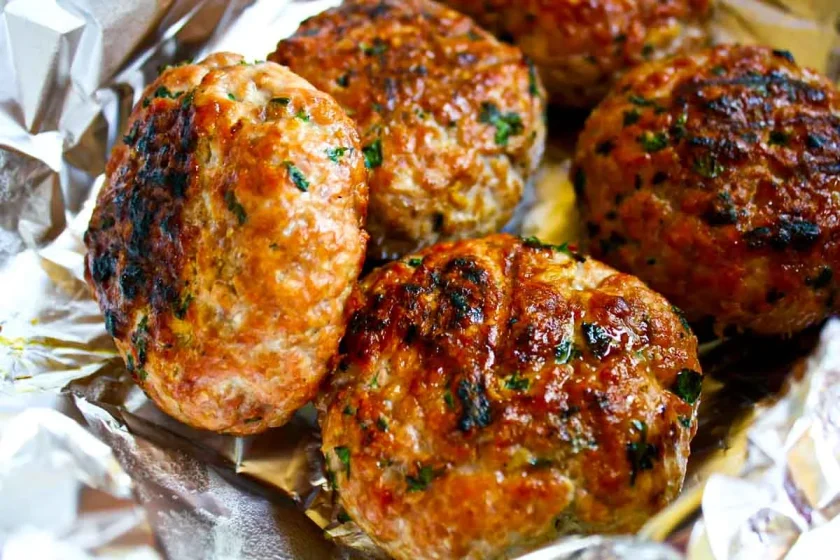 Cinco hamburguesas de carne dorada a la parrilla con hierbas descansan sobre papel de aluminio arrugado.