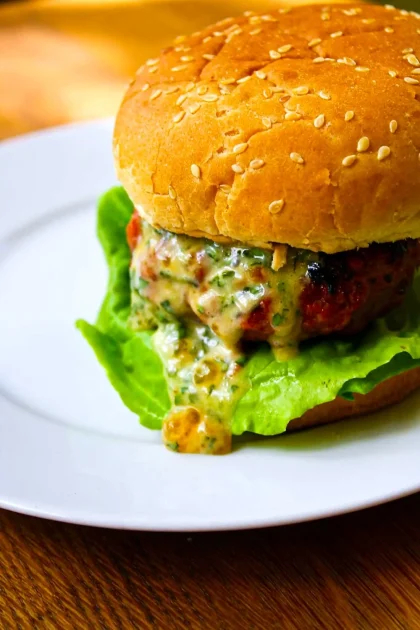 A burger with lettuce and a sesame seed bun, featuring apricot-chipotle mayo dripping from the patty, served on a white plate.