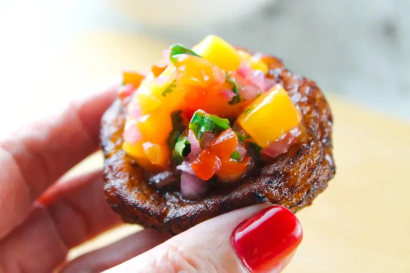 A hand with red-painted nails holding a small fried plantain cup topped with diced mango, red bell pepper, onion, and cilantro.