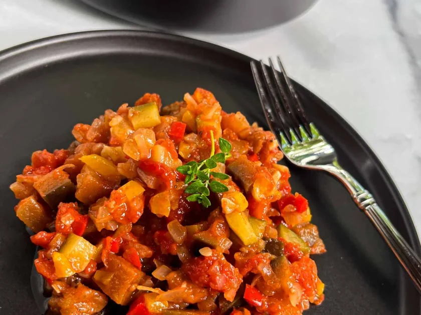 Una ración de ratatouille adornada con un ramito de tomillos fresco, en un plato negro, junto a un tenedor.