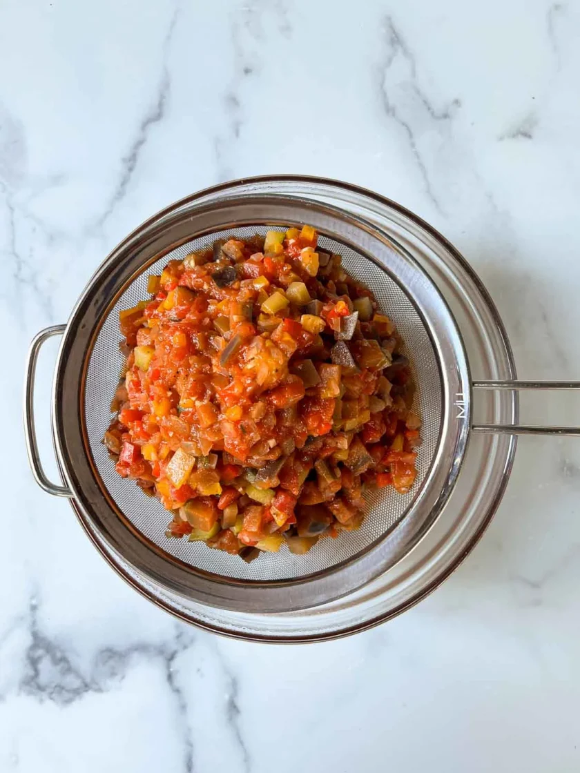 A metal strainer filled with diced cooked vegetables, placed over a glass bowl on a white marble surface.