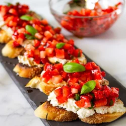 Rebanadas de pan tostado cubiertas con ricotta, fresas troceadas y hojas de albahaca fresca dispuestas en una bandeja negra; al fondo, un cuenco con la mezcla de fresas.