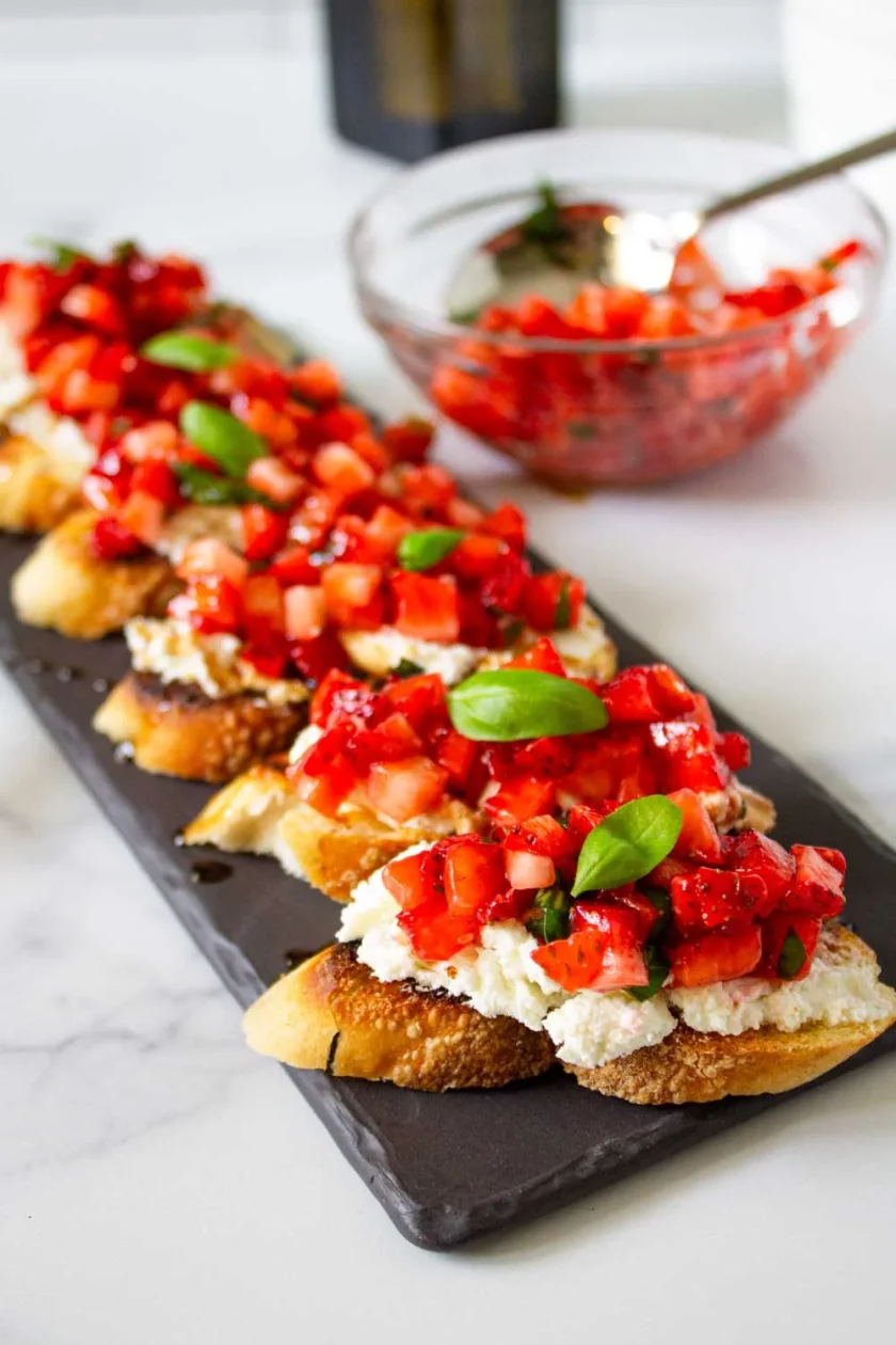 Rebanadas de pan tostado cubiertas con queso de cabra, fresas cortadas en cubitos, hojas de albahaca y glaseado balsámico, dispuestas en una tabla de pizarra , para servir con un cuenco de mezcla de fresas al fondo.