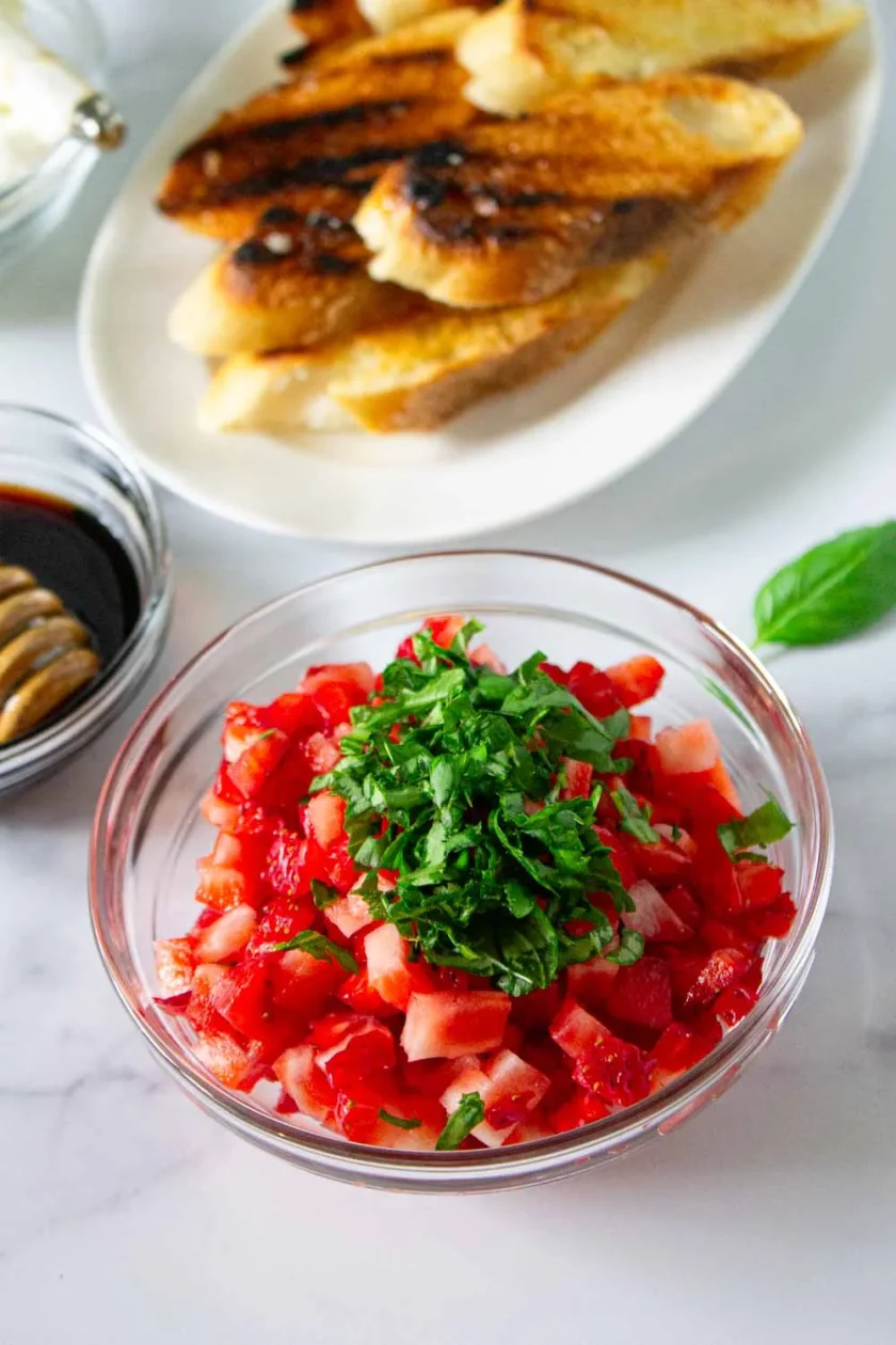 Un cuenco de cristal con fresas picadas y albahaca fresca descansa sobre una superficie blanca, con rebanadas de pan tostado y un cuenco con reducción de vinagre balsámico al fondo.