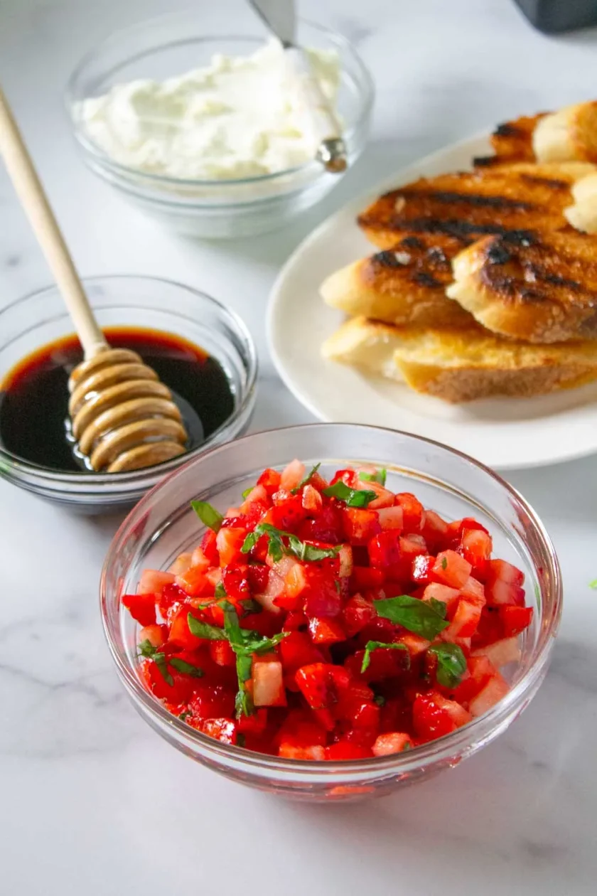 Un cuenco de cobertura de crostini de fresa, rebanadas de pan tostado, un cuenco de reducción de balsåmico y un cuenco de queso de cabra fresco, todo sobre una superficie de mármol.