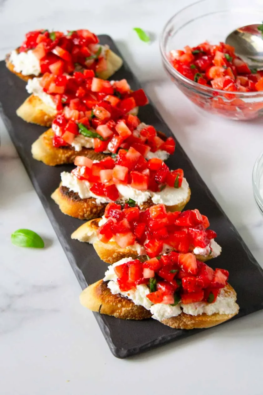Plato rectangular de pizarra con rebanadas de pan tostado cubiertas con queso de cabra  y mezcla de fresas en dados, con cuencos de mezcla de fresas picadas al fondo.