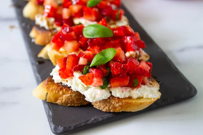Rebanadas de pan tostado cubiertas con queso de cabra, fresas cortadas en dados y hojas de albahaca fresca, dispuestas en una bandeja oscura para servir.