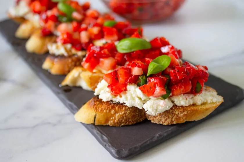 Rebanadas de pan tostado cubiertas con queso de cabra, dfresas cortaditas y hojas de albahaca, dispuestas en una bandeja oscura para servir.