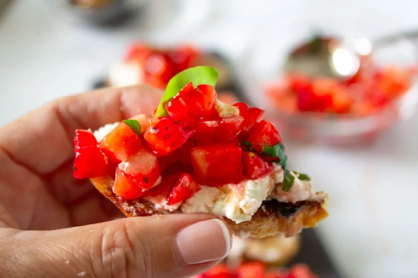 Una mano sostiene un crostini con queso de cabra fresco, fresas troceadas y una hoja de albahaca.