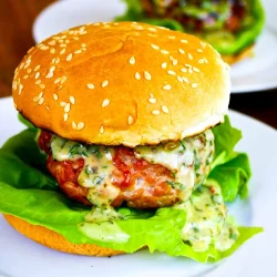 Una hamburguesa con lechuga y salsa cremosa en un pan con semillas de sésamo, servida en un plato blanco, con otra hamburguesa al fondo.