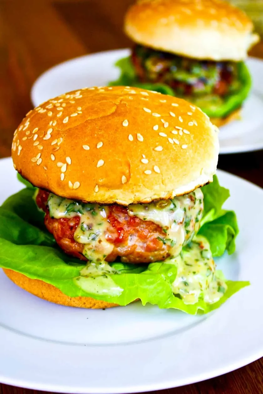 Una hamburguesa con lechuga y salsa cremosa en un pan con semillas de sésamo, servida en un plato blanco, con otra hamburguesa al fondo.