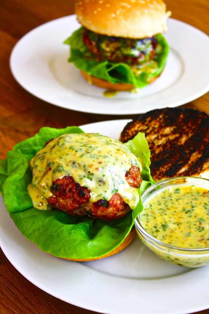 A burger patty with apricot chipotle mayo on lettuce and a toasted bun, served with extra sauce in a small bowl; another assembled burger in the background.