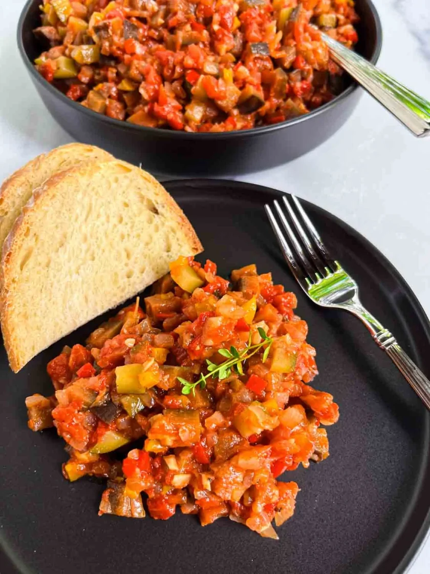 Una ración de ratatouille un plato negro con dos rebanadas de pan y un tenedor; hay más ratatouille en un cuenco al fondo.