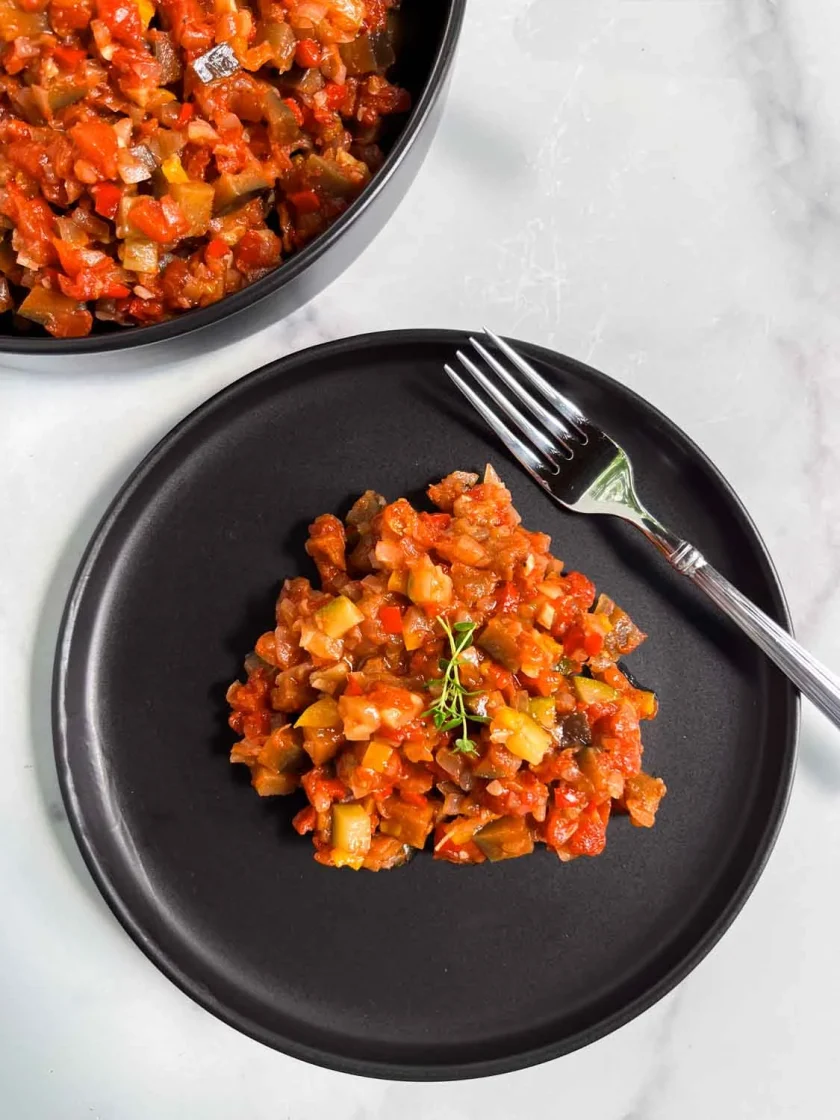 A serving of diced vegetable ratatouille on a black plate, garnished with fresh thyme, next to a fork on a white surface.