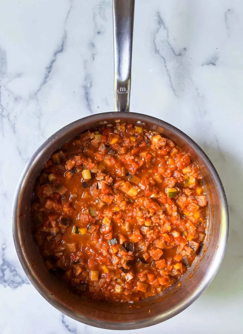 A stainless steel pan filled with a chunky tomato-based ratatouille sits on a white marble surface.