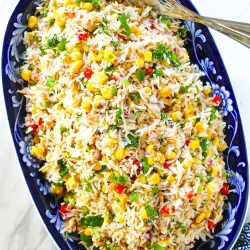 Plato ovalado de color azul relleno de ensalada de arroz y atún con maíz, cilantro y verduras rojas y verdes cortadas en dados, con una cuchara para servir al lado.