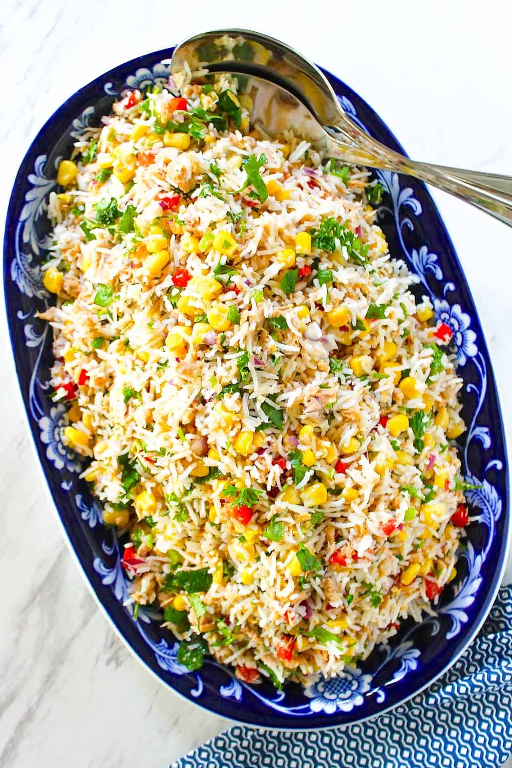 Plato ovalado de color azul relleno de ensalada de arroz y atún con maíz, cilantro y verduras rojas y verdes cortadas en dados, con una cuchara para servir al lado.