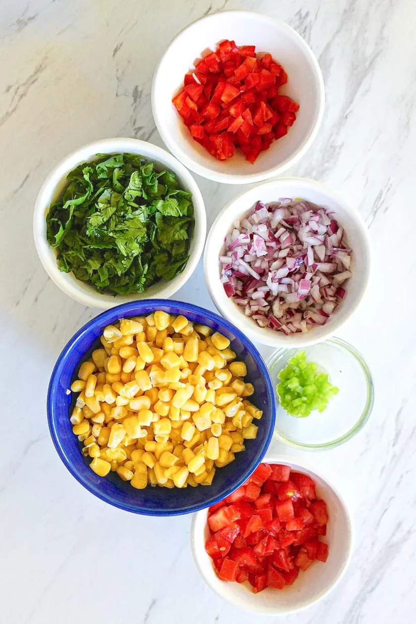 Seis cuencos con pimiento rojo cortado en dados, cilantro picado, cebolla roja picada, pimiento verde picado, tomate cortado en dados y granos de maíz dispuestos sobre una superficie blanca.