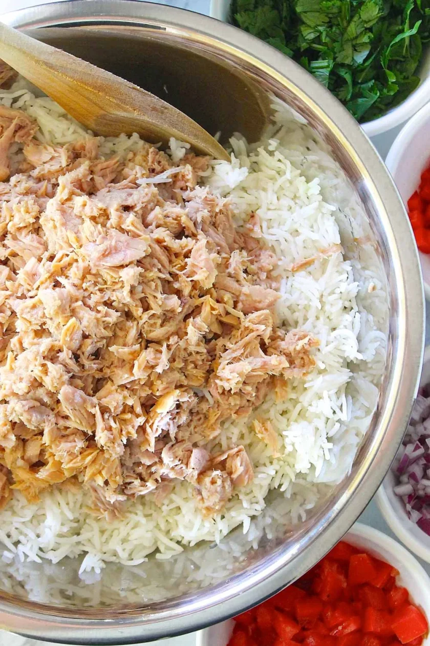 Un cuenco de metal lleno de arroz blanco cocido cubierto de atún desmenuzado, rodeado de pequeños cuencos de verduras y hierbas picadas.