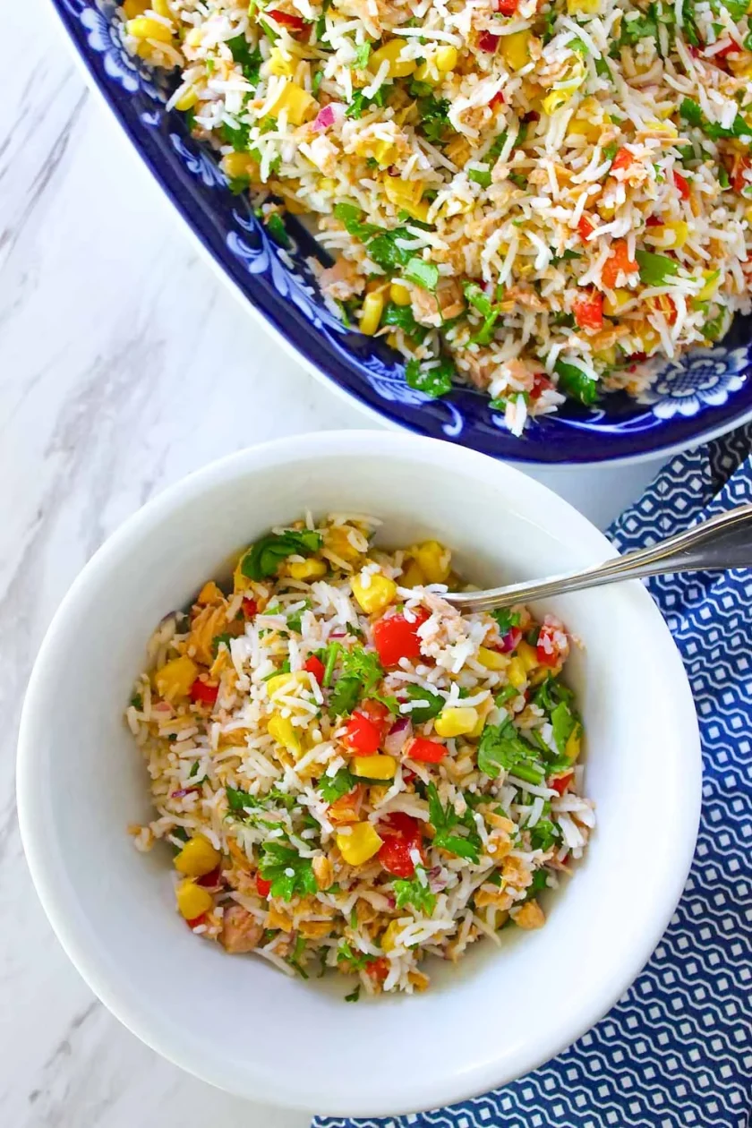 Un cuenco blanco de ensalada de arroz con atúb y maíz, pimientos rojos, hierbas y verduras se encuentra junto a una fuente más grande de la misma ensalada sobre una superficie de mármol.