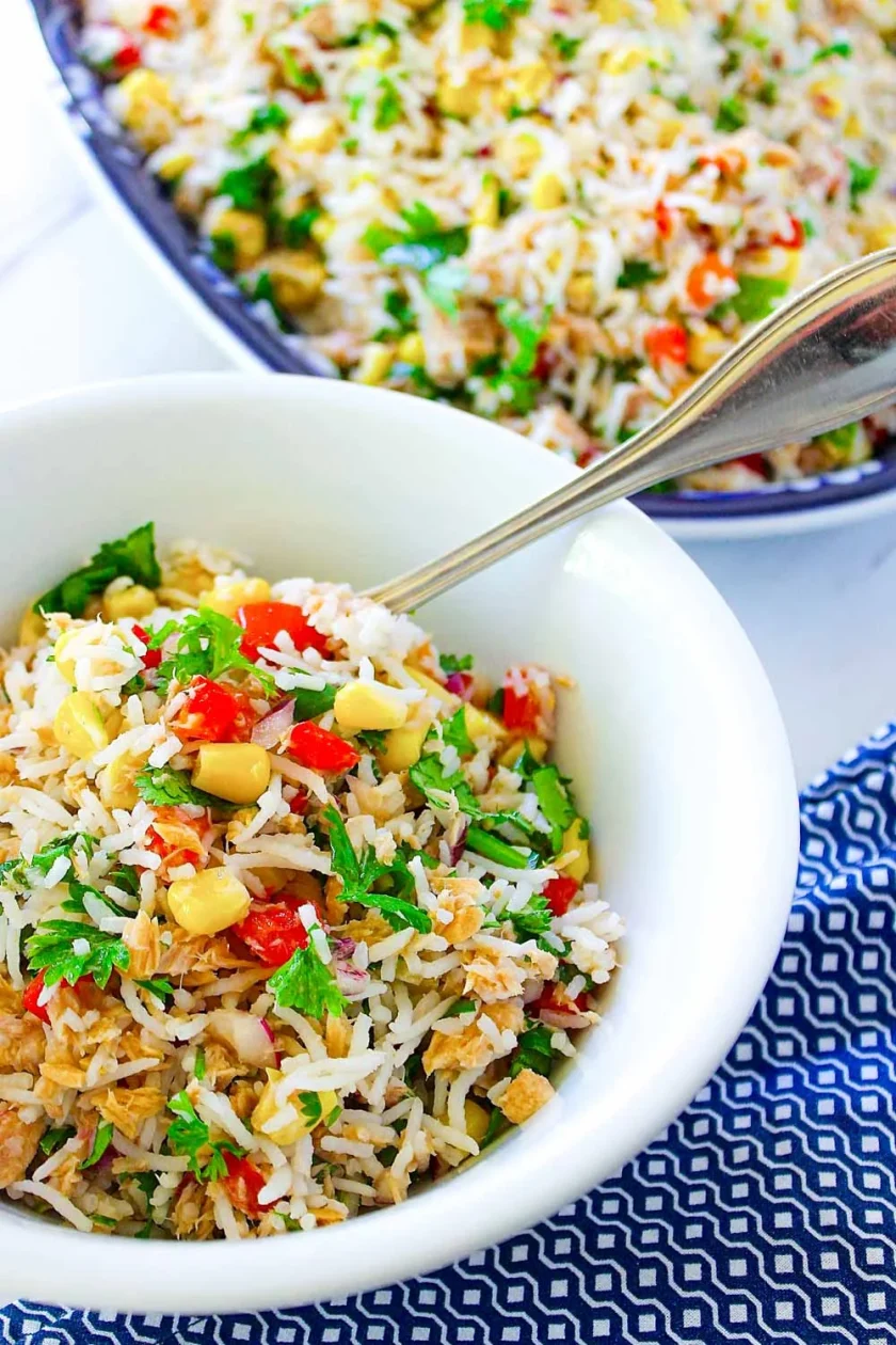 Un cuenco blanco lleno de ensalada de arroz mezclada con atún, maíz, pimientos rojos, hierbas y un tenedor, con un cuenco más grande de la misma ensalada al fondo.
