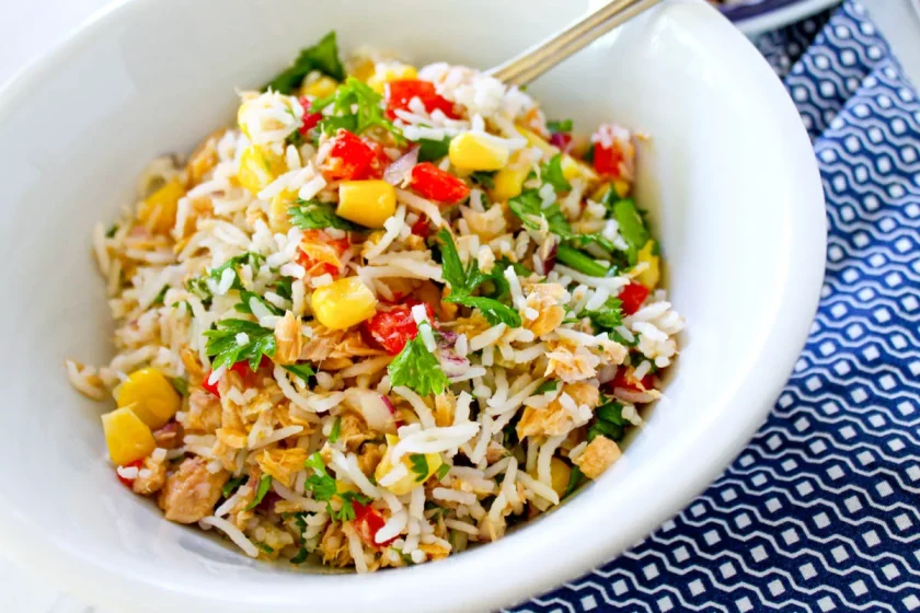 Un bol de ensalada de arroz con atün, maíz, pimientos rojos, hierbas picadas y trozos de pollo, servido en un plato blanco con cuchara.