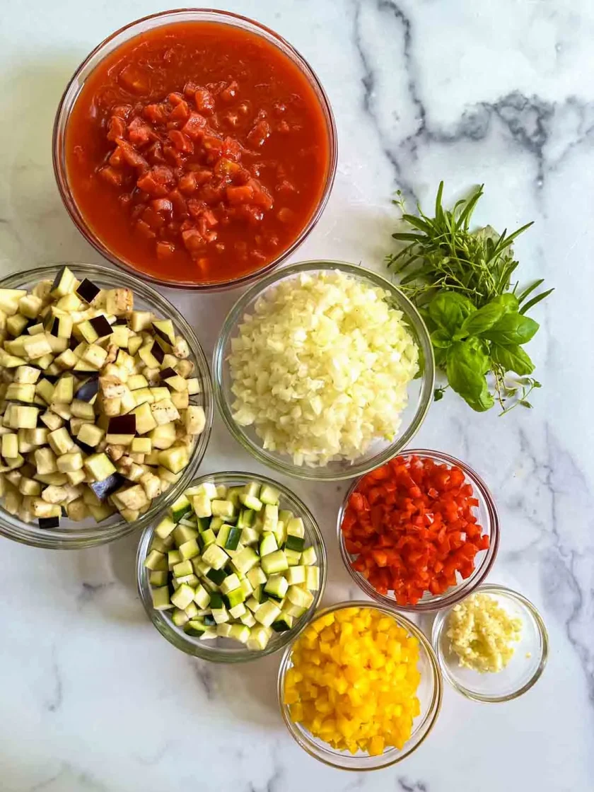 Bowls of chopped vegetables—eggplant, zucchini, yellow and red bell peppers, onion, garlic, and tomatoes—plus fresh herbs on a marble surface.