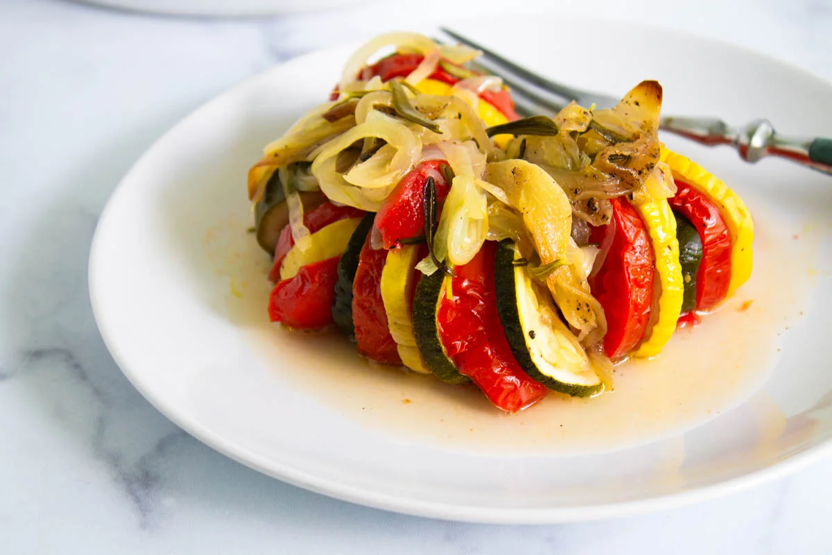 Sliced tomatoes, zucchini, and yellow squash topped with cooked onions, arranged on a white plate with a fork.