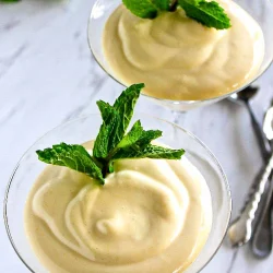 Dos copas de cristal llenas de un postre cremoso, cada una adornada con ramitas de menta fresca, colocadas sobre una superficie blanca con cubiertos y hojas de menta al fondo.