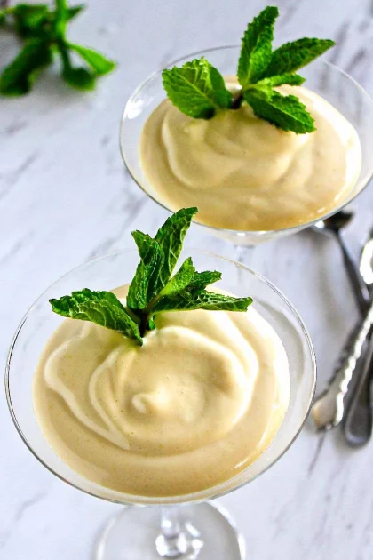 Dos copas de cristal llenas de un postre cremoso, cada una adornada con ramitas de menta fresca, colocadas sobre una superficie blanca con cubiertos y hojas de menta al fondo.
