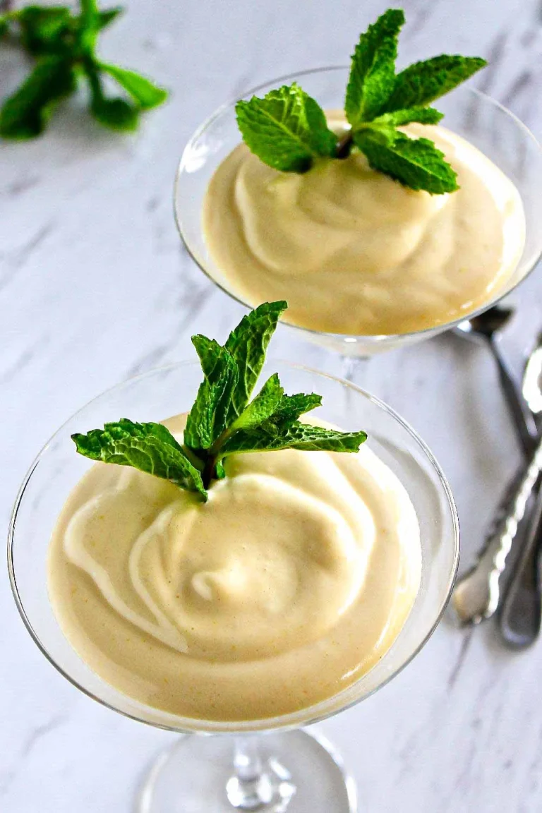 Dos copas de cristal llenas de un postre cremoso, cada una adornada con ramitas de menta fresca, colocadas sobre una superficie blanca con cubiertos y hojas de menta al fondo.
