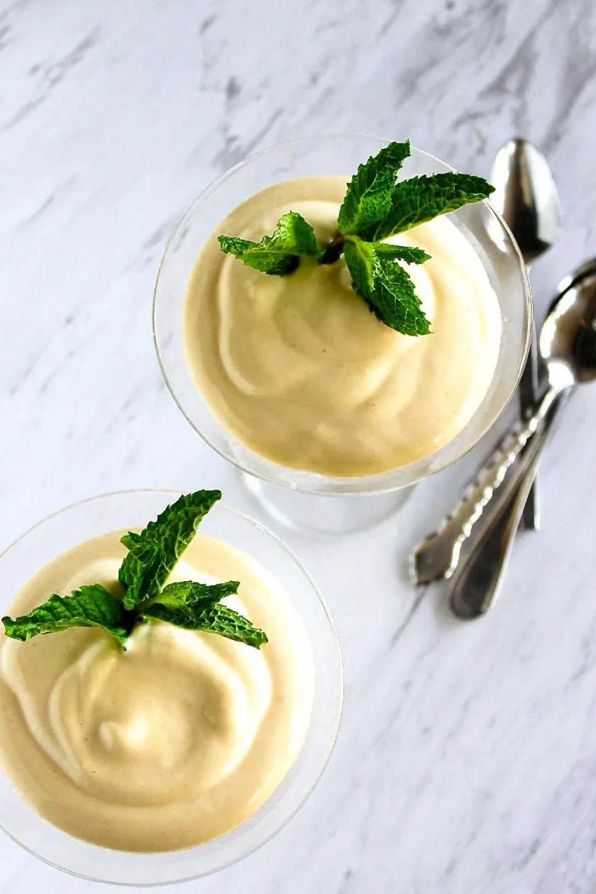 Dos copas de postre cremoso adornadas con hojas de menta fresca, colocadas sobre una superficie de mármol blanco junto a un par de cucharas de plata.