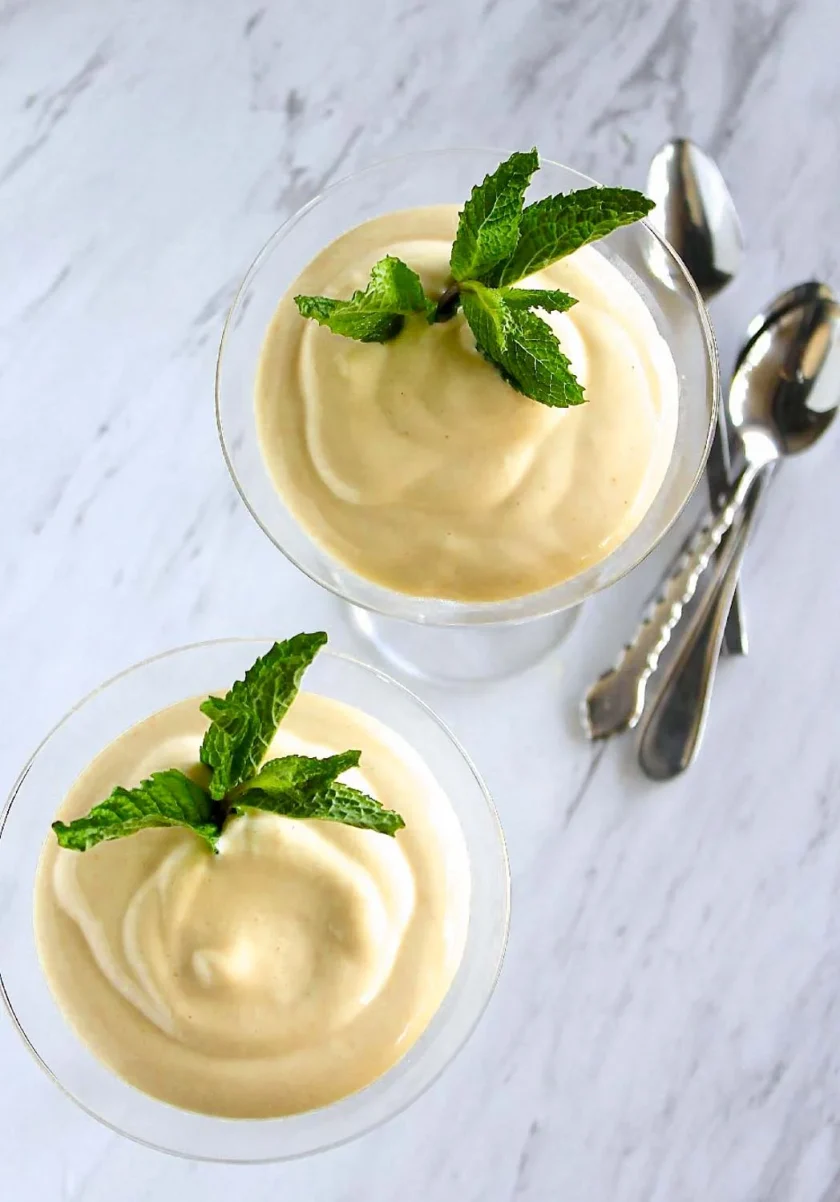 Dos copas de mousse cremosa adornadas con hojas de menta fresca, colocadas sobre una superficie de mármol con tres cucharas apiladas junto a ellas.
