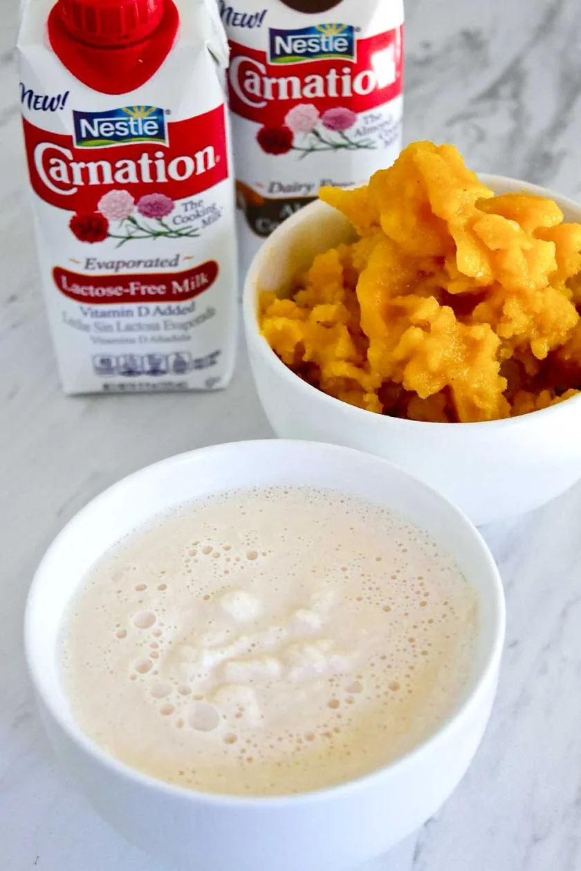 Una taza de una bebida cremosa, un cuenco de granizado de marchita, y dos envases de leche evaporada sin lactosa «Nestlé Carnation» sobre una superficie de mármol.