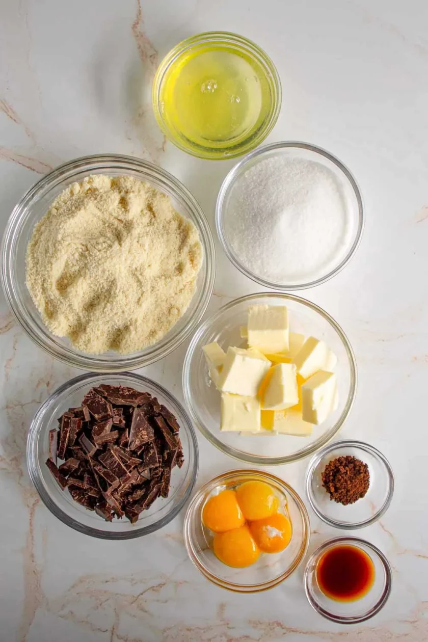 Siete cuencos de cristal con yemas y claras de huevo separadas, azúcar, mantequilla en dados, extracto de vainilla, café instantáneo, chocolate picado y harina de almendra sobre una superficie de mármol.